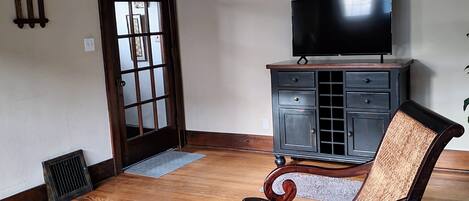 A living room with a wooden floor, a patterned rug, a dark wooden chair, a television on a cabinet, and a door with frosted glass panels.