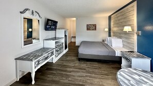 A hotel room with a bed, a desk with drawers, a mirror, a TV, and a wall-mounted lamp.