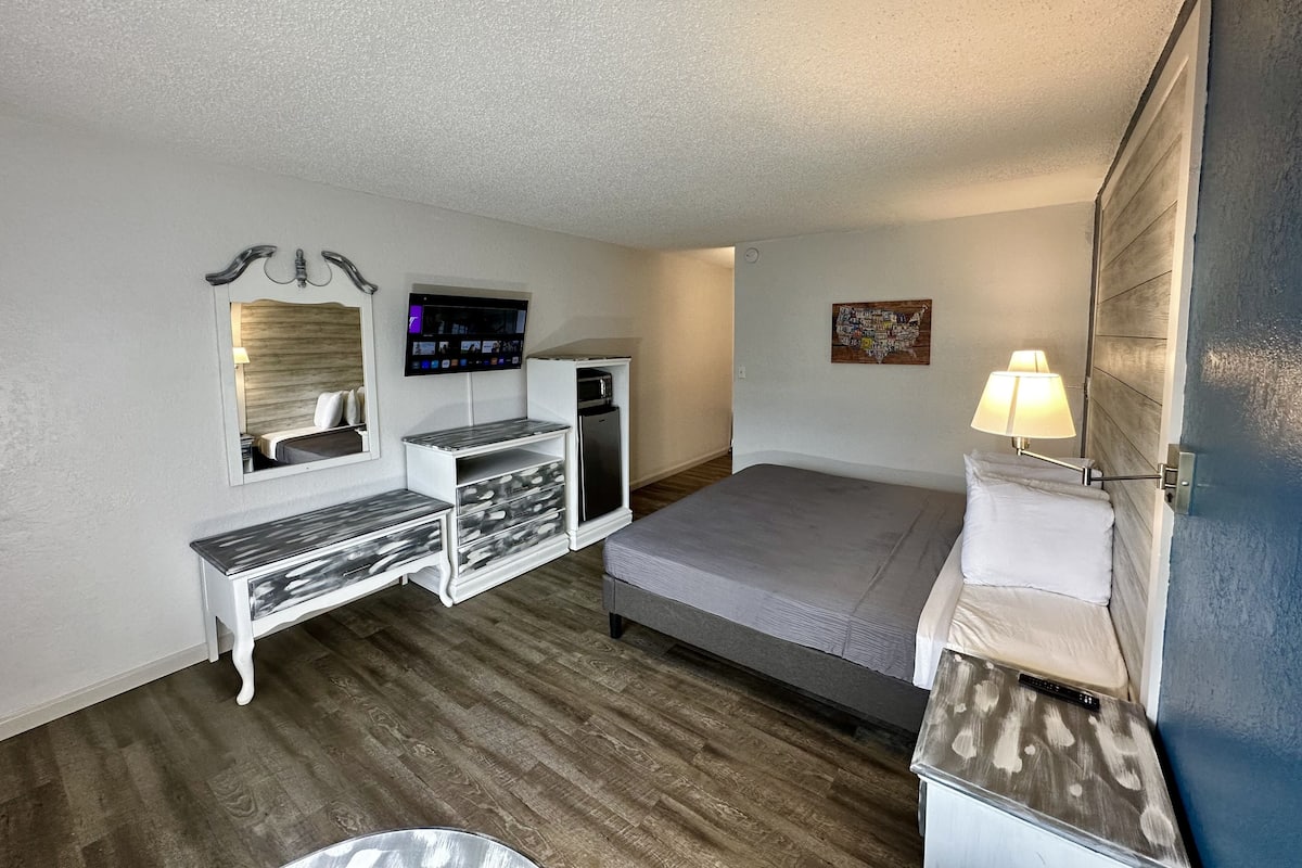 A compact hotel room with a bed, a desk with drawers, a small refrigerator, a flat-screen TV, and a wall-mounted lamp.