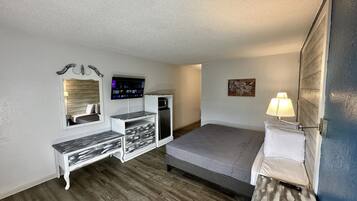 A compact hotel room with a bed, a desk with drawers, a small refrigerator, a flat-screen TV, and a wall-mounted lamp.