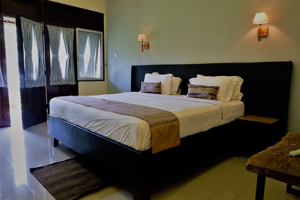 Uma cama bem arrumada com lençóis brancos e um manta marrom, duas mesas de cabeceira com abajures e uma janela com cortinas.