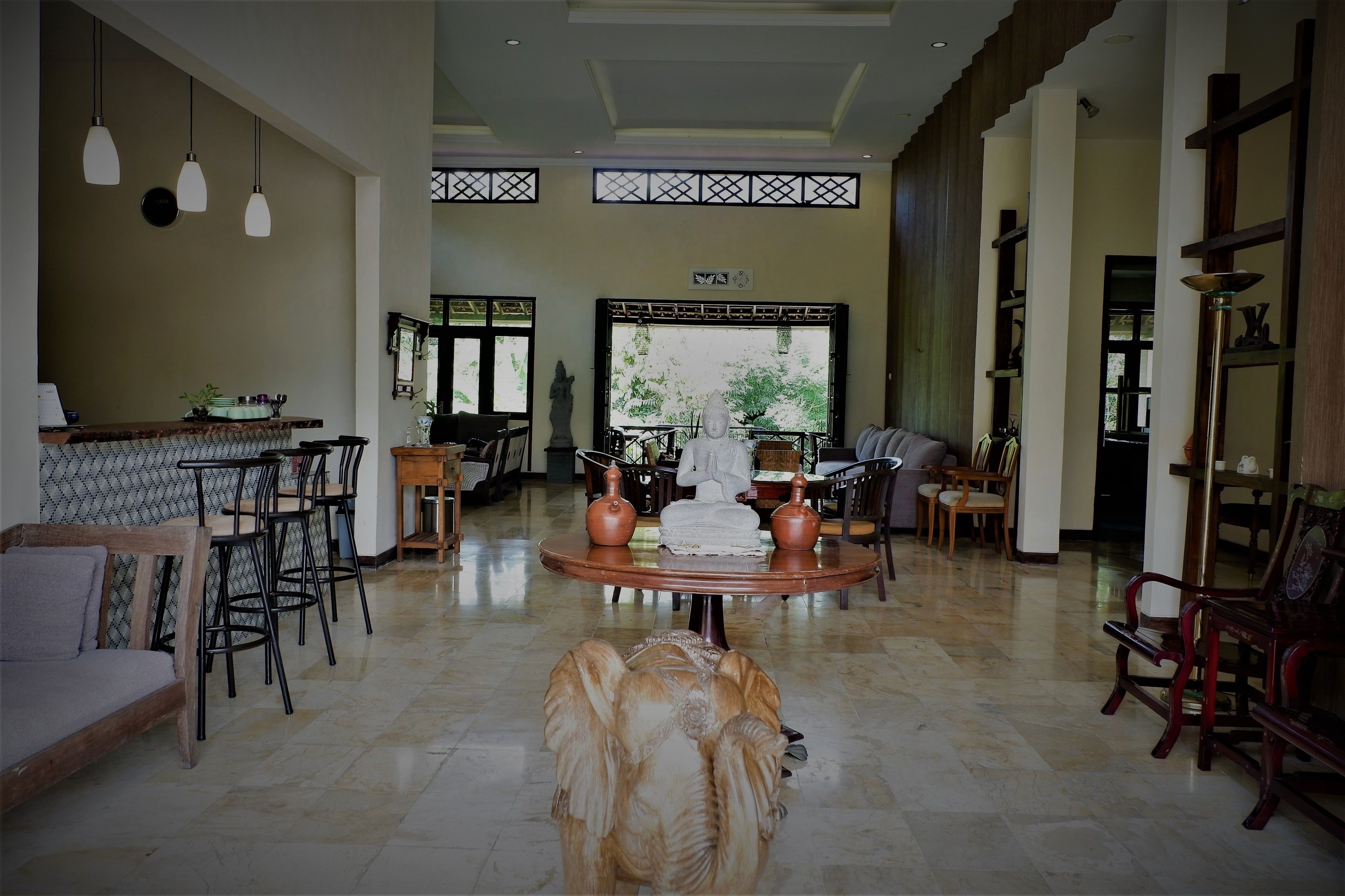 A spacious living area with a high ceiling, a central round table with two vases, and a large window with a view of greenery.