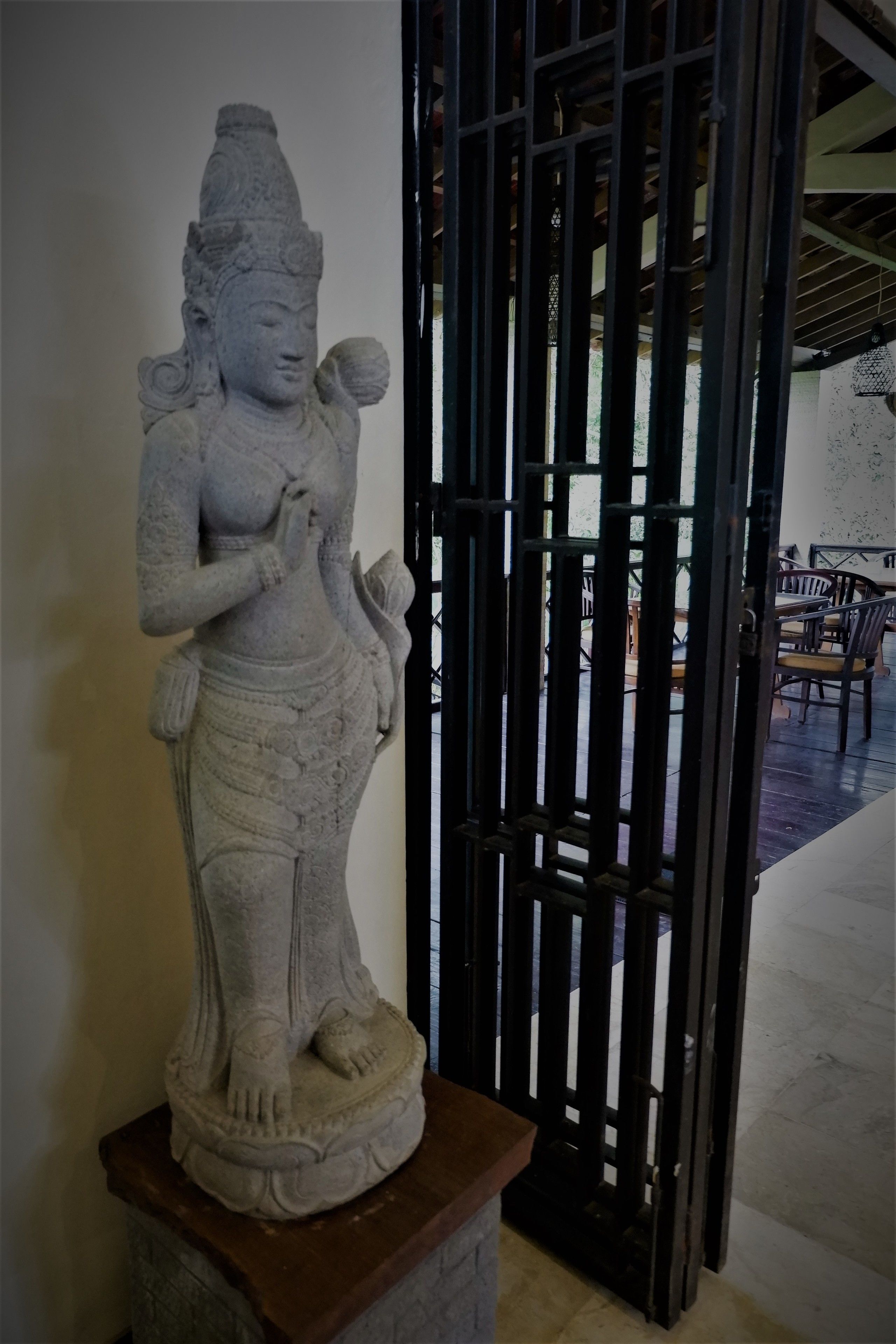 Una estatua de piedra de una deidad con intrincados grabados, ubicada sobre un pedestal en una habitación con puerta de madera y suelo de baldosas.