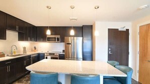 Una cocina moderna con armarios oscuros, electrodomésticos de acero inoxidable, una isla central con taburetes y luminarias colgantes, además de una puerta de madera.