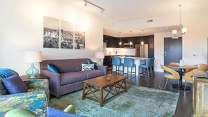 Una sala de estar moderna con un sofá, una mesa de centro y sillas. Cuenta con una zona de cocina con taburetes de bar y una mesa de comedor.