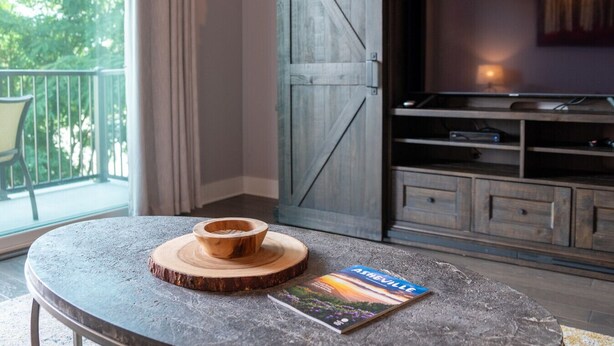 Ein Zimmer mit einem Steintisch, einer Holzschüssel und einer Zeitschrift. Ein Fernseher steht auf einem Holzgestell und es gibt eine große Schiebetür im Scheunenstil.