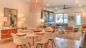 A modern dining area with a wooden table set for four, surrounded by white chairs. A large pendant light hangs above the table, and the room features a mix of contemporary and classic furniture. - 243 Short Coxe Ave Townhome (Asheville)