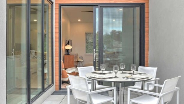 A modern dining area with a glass table and white chairs, a wooden wall, and a sliding glass door leading to a room with a lamp and a sofa. - 243 Short Coxe Ave Townhome (Asheville)