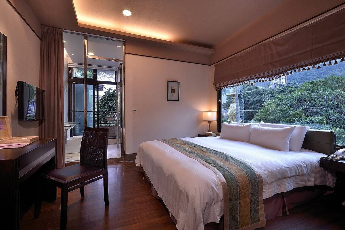 Une chambre d’hôtel avec un lit, un bureau, une chaise, une fenêtre donnant sur un espace verdoyant et un tableau encadré au mur.
