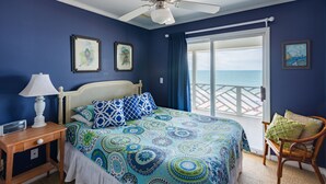 Quarto com cama, mesa de cabeceira, cadeira, ventilador de teto e vista para o oceano através de uma porta de vidro deslizante.