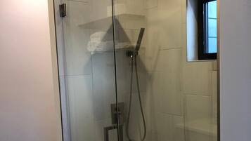 Una doccia moderna, completamente vetrata, con una doccetta manuale e una soffione fisso, uno scaffale piccolo e una finestra sulla parete destra.