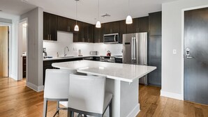 A modern kitchen with dark cabinets, stainless steel appliances, a central island with bar stools, pendant lighting, and a wooden floor. - Roomy and Accessible Downtown Balcony Condo (Asheville)