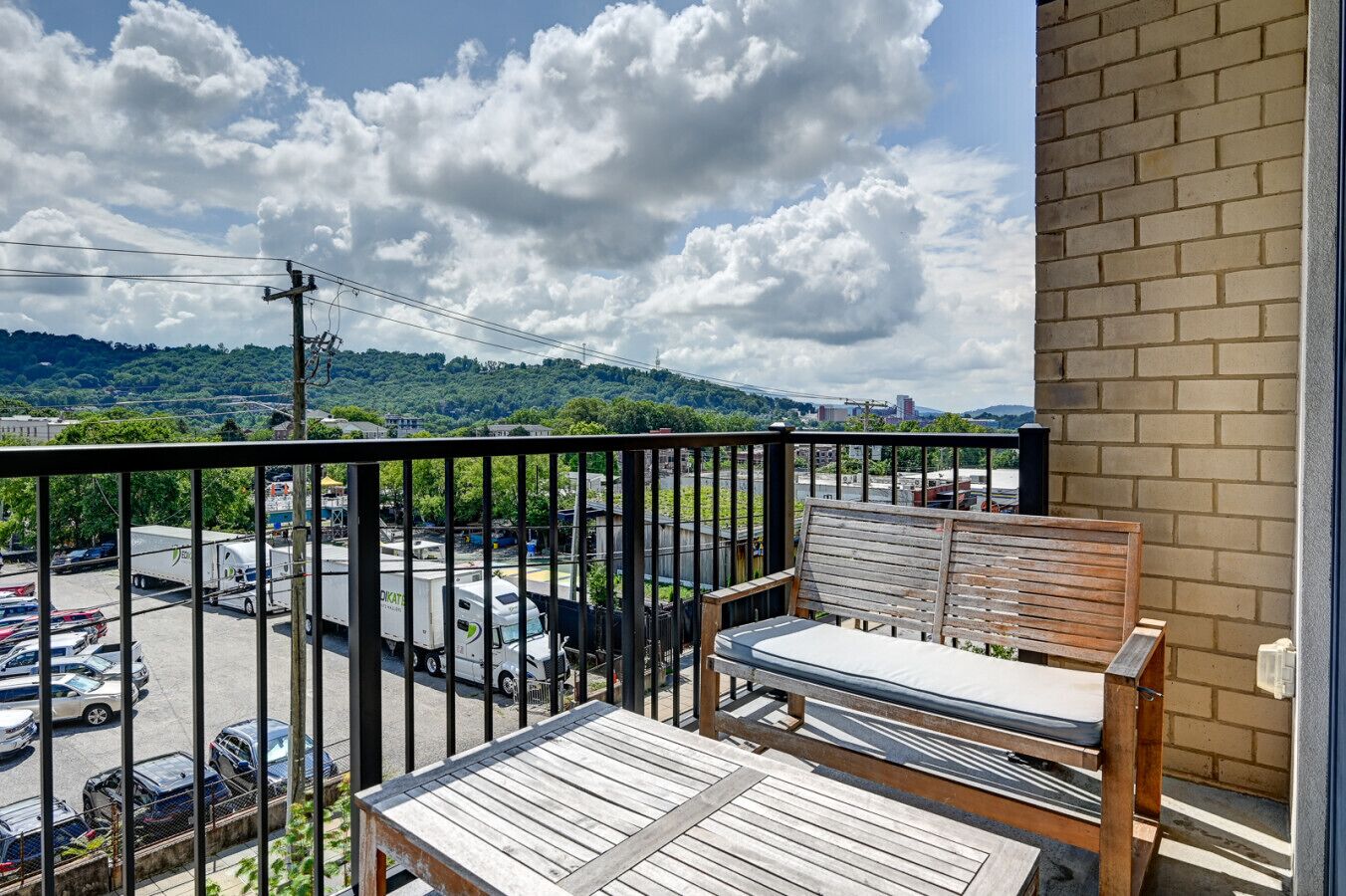 Un balcon avec un banc et une table en bois, donnant sur un parking et sur un paysage urbain composé d’immeubles et d’espaces verts.