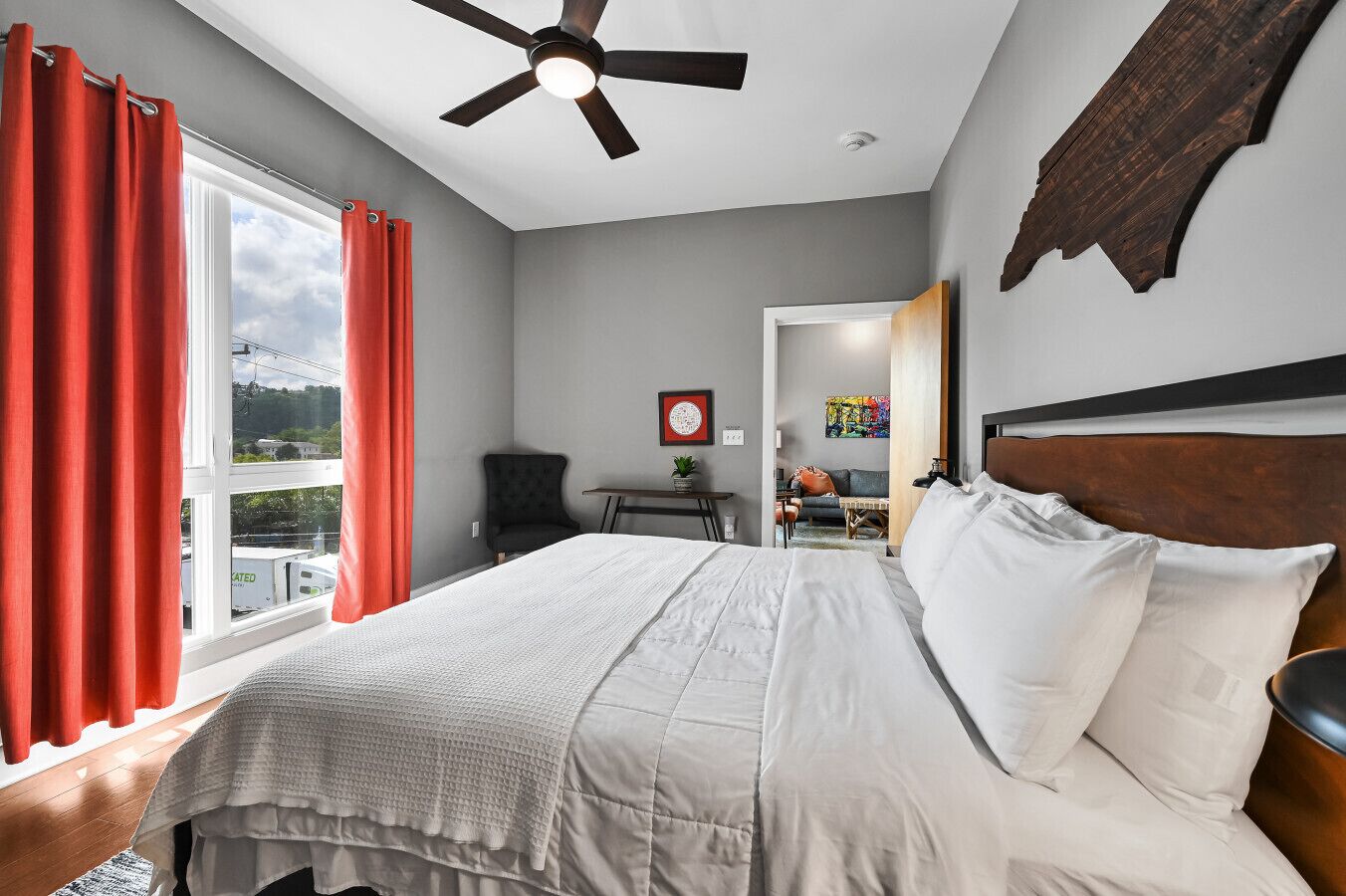 A bedroom with a bed, a ceiling fan, a window with red curtains, and a view of the outside.