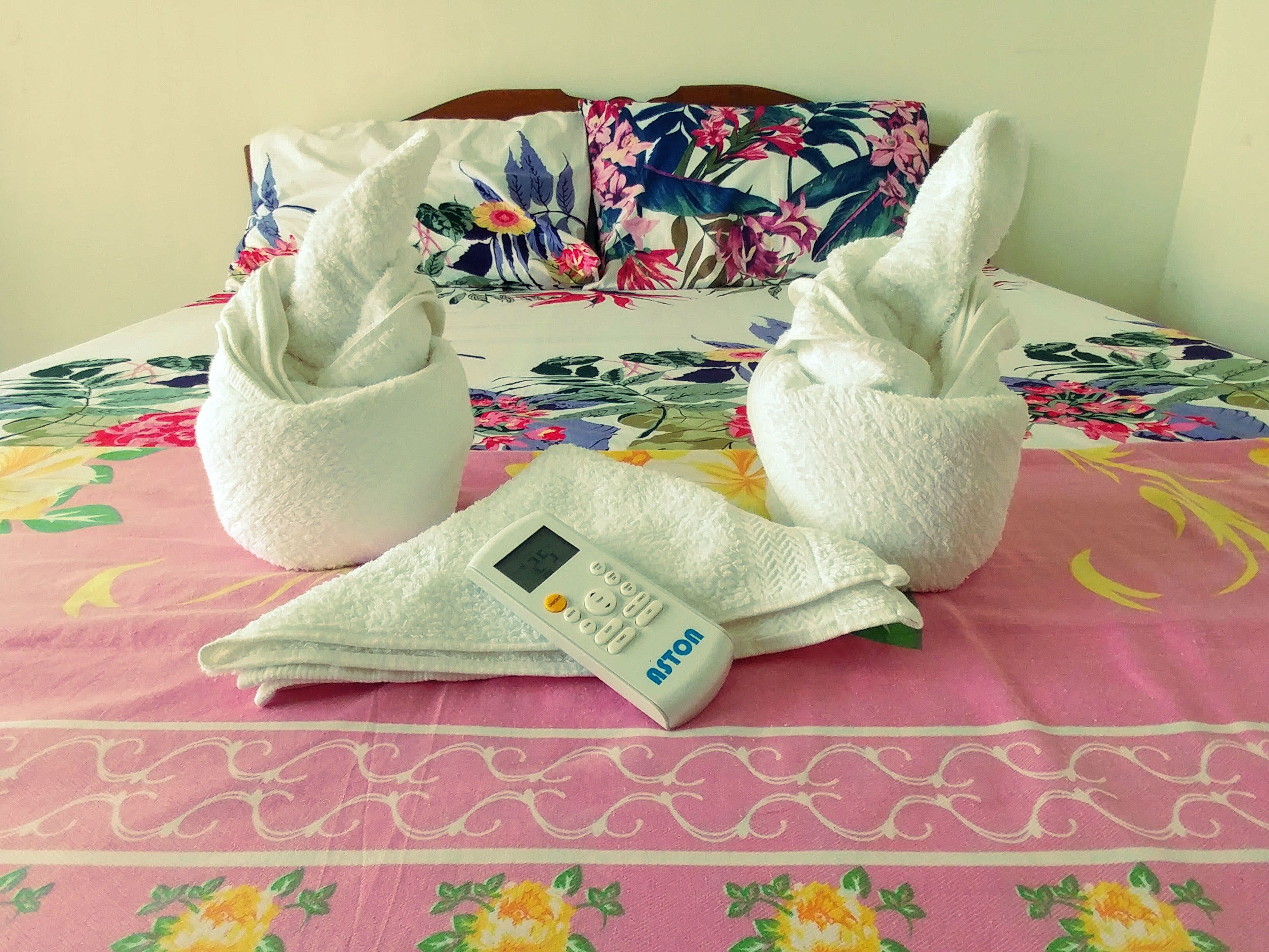 A bed with floral bedding, two white towels folded into swans, and a digital clock displaying 27.