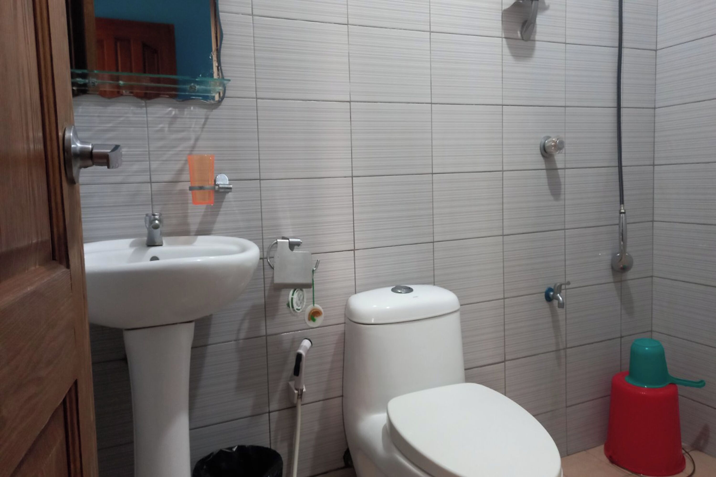 A bathroom with a white sink, toilet, and shower. There is a red trash bin and a glass shelf above the sink.