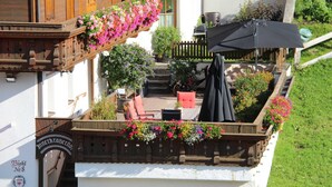 Un balcón de madera con cestas de flores colgantes, mobiliario exterior y una zona de barbacoa.