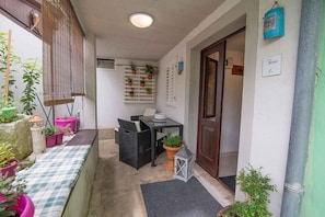 A narrow outdoor corridor with a checkered bench, wicker furniture, potted plants, and a lantern.