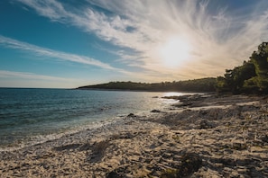A rocky shoreline with clear water, a distant tree-covered hill, and a bright sun in the sky.