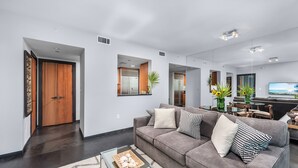 Una sala de estar moderna con un sofá gris, una mesa de centro de vidrio y un televisor. Hay una zona de comedor con una mesa y sillas, y una cocina al fondo.