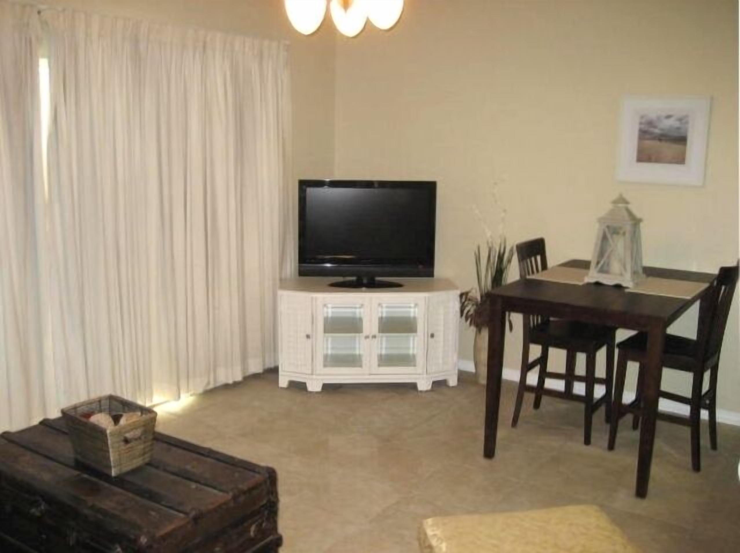 A living room with a television on a cabinet, a dining table with chairs, a wooden bench, and a window with curtains.