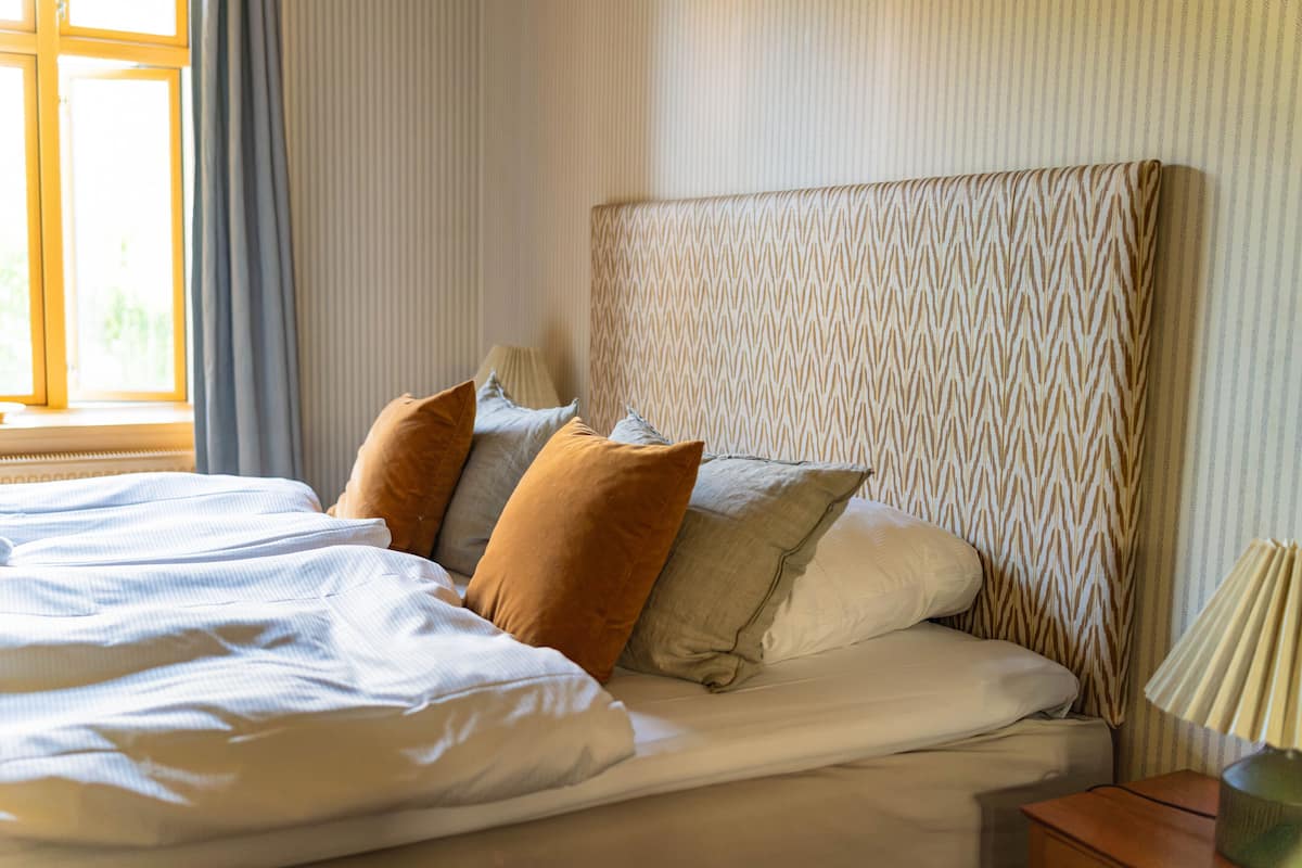A neatly made bed with white linens and several pillows, a bedside lamp, and a window with curtains.
