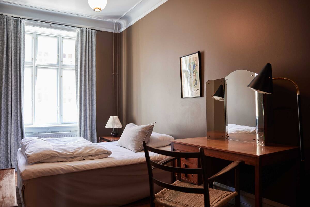 A hotel room with a bed, a desk, a chair, a lamp, a picture on the wall, and a window with curtains.