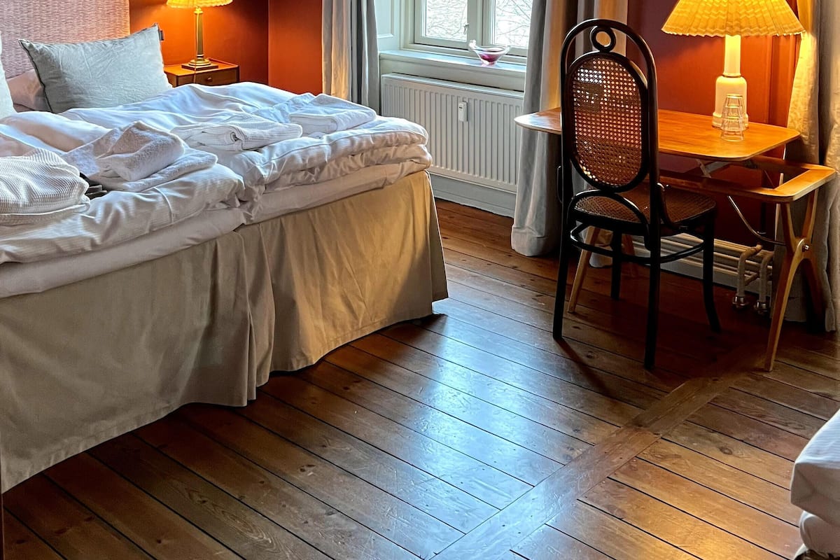 A hotel room with a bed, two bedside lamps, a chair, a table, and a window with curtains.