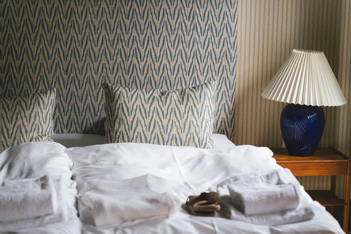 A neatly made bed with white linens, a blue vase on a wooden nightstand, and a patterned wall in the background.