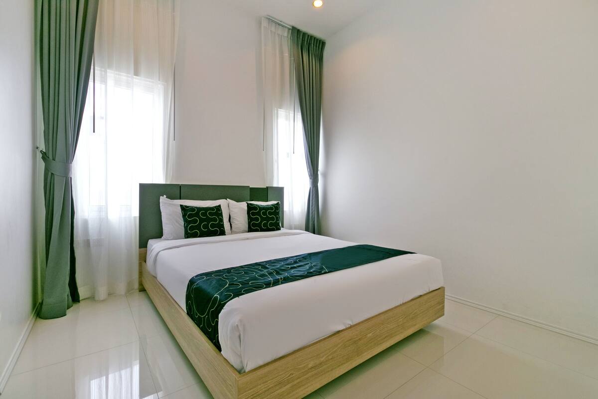 A neatly made bed with a wooden frame and green bedding, positioned in a room with white walls and a large window with sheer curtains.