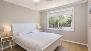 A bedroom with a bed, two bedside tables with lamps, a window with a view of trees, and a ceiling fan. - SEA BREEZE - Beachside Family Haven (Broadwater)