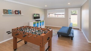 A room with a foosball table, a television, a sofa, and a window with a view of a backyard. - SEA BREEZE - Beachside Family Haven (Broadwater)