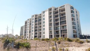 A multi-story residential building with balconies, located near a sandy area with sparse vegetation.