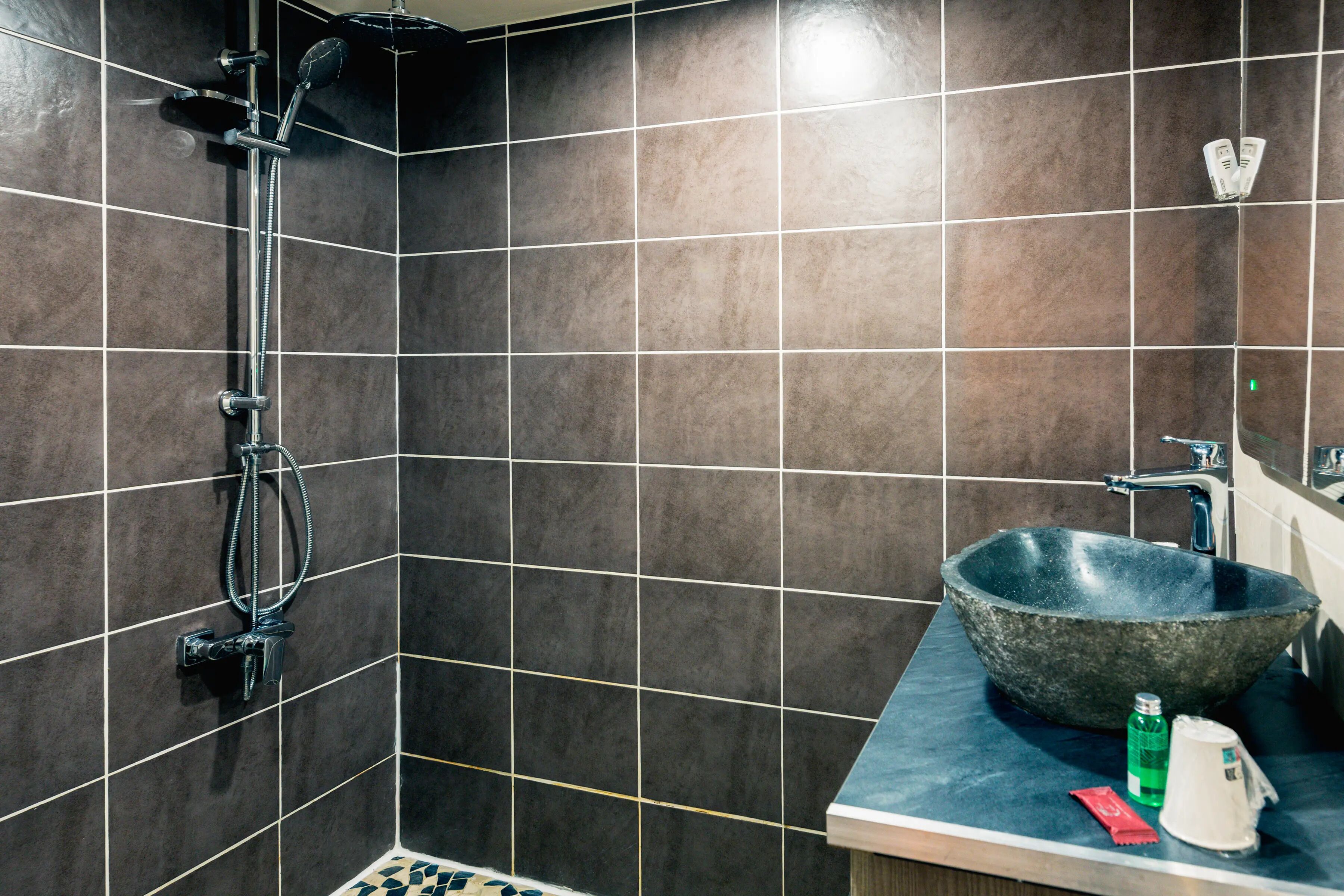 Ein Badezimmer mit dunkel gefliesten Wänden, einer Dusche mit Duschhandstück, einem großzügigen Steinklopfbac und einer Ablagefläche mit Seifenspender und Flasche.
