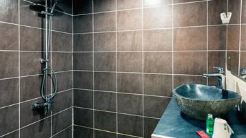 A bathroom with dark tiled walls, a shower with a handheld showerhead, a large stone sink, and a countertop with a soap dispenser and a bottle.