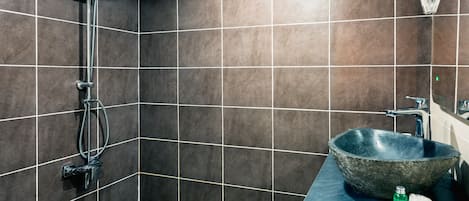 A bathroom with dark tiled walls, a shower with a handheld showerhead, a large stone sink, and a countertop with a soap dispenser and a bottle.