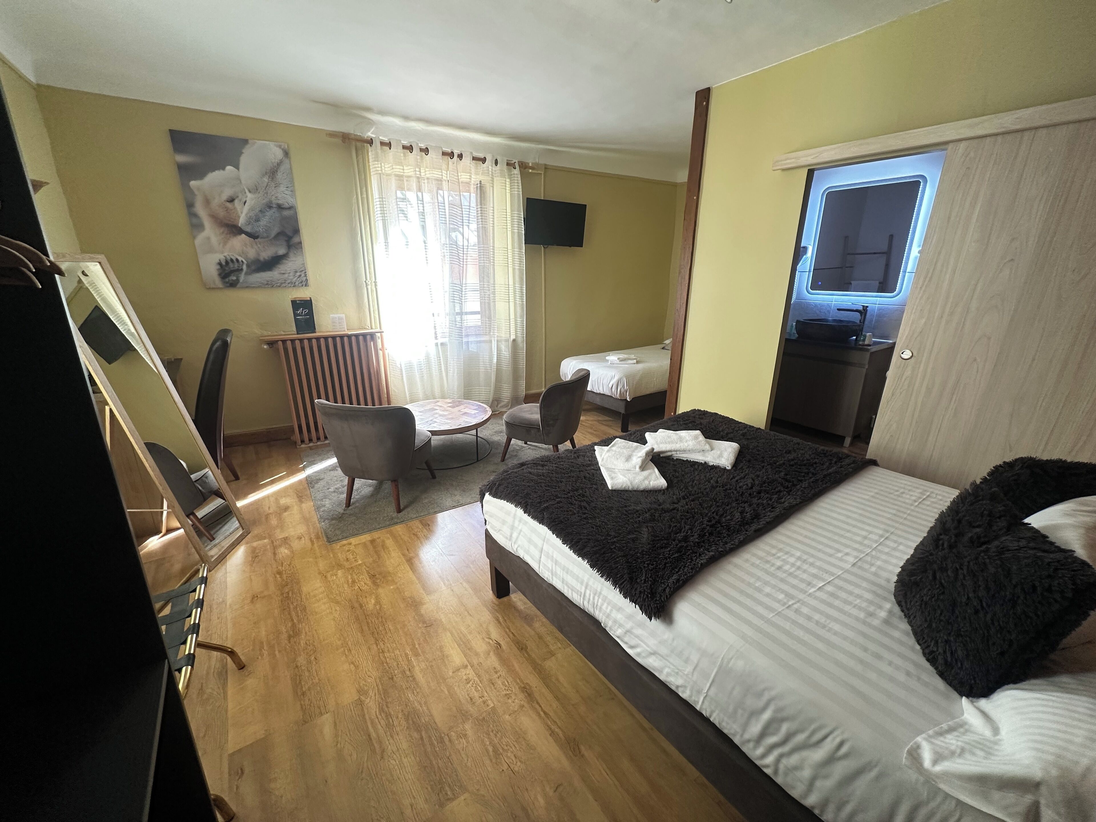 A hotel room with a bed, a desk, a chair, a small table, a window, and a painting on the wall.