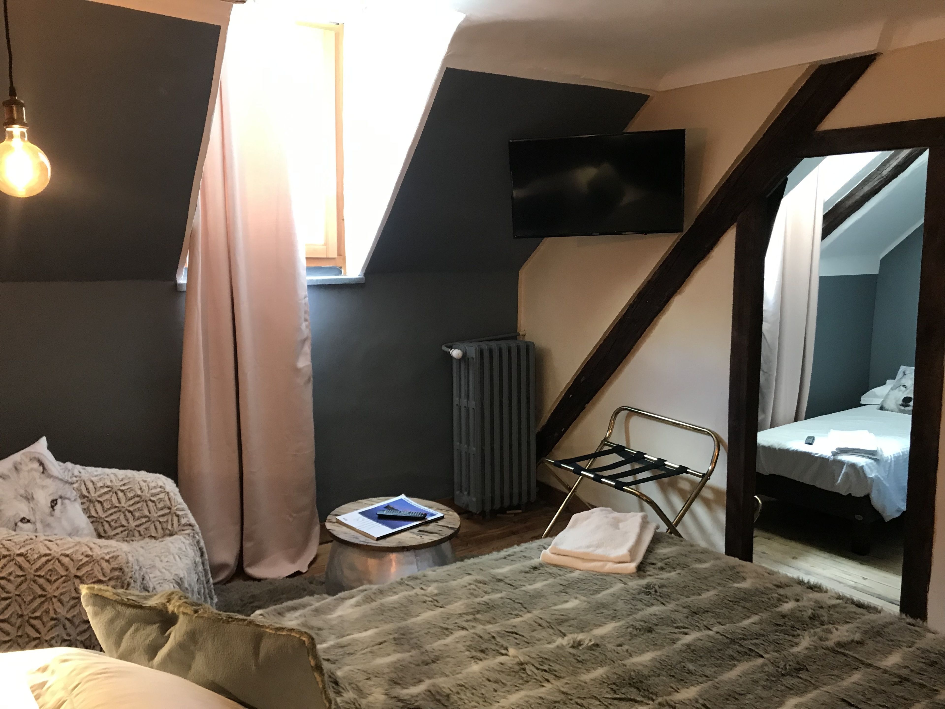 Un dormitorio compacto con techo inclinado, una cama con colcha blanca, una mesita de noche pequeña con un libro y un control remoto, un radiador y un televisor montado en la pared.