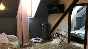 Un dormitorio compacto con techo inclinado, una cama con colcha blanca, una mesita de noche pequeña con un libro y un control remoto, un radiador y un televisor montado en la pared.