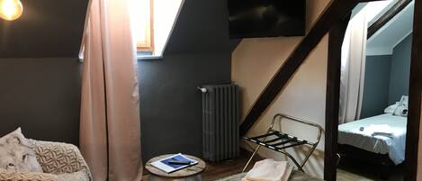 Un dormitorio compacto con techo inclinado, una cama con colcha blanca, una mesita de noche pequeña con un libro y un control remoto, un radiador y un televisor montado en la pared.