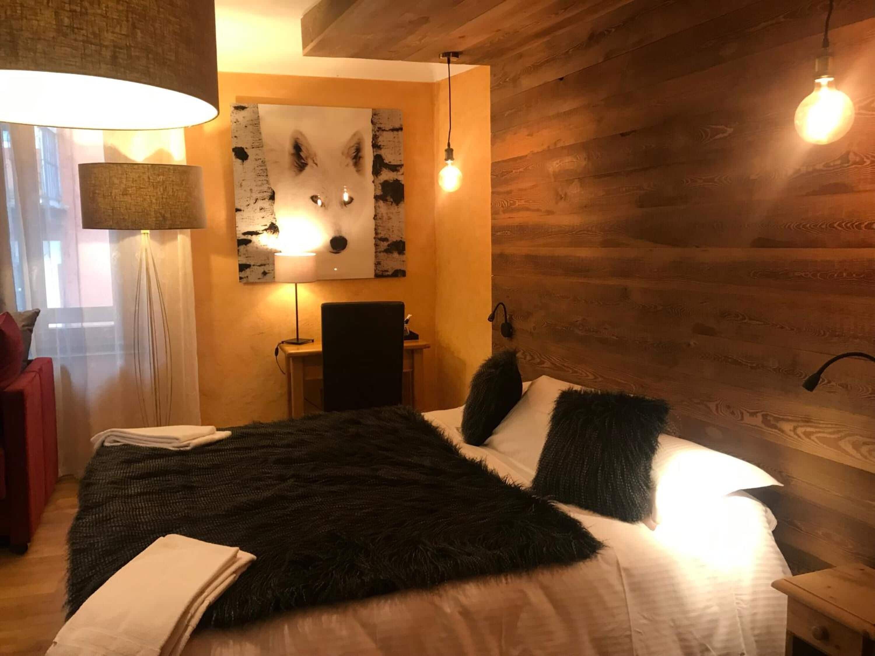 A cozy bedroom with a bed, a desk, and a chair. There is a lamp on the desk and a painting on the wall.