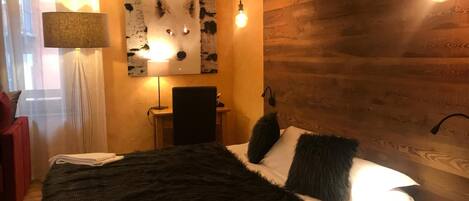 A cozy bedroom with a bed, a desk, and a chair. There is a lamp on the desk and a painting on the wall.
