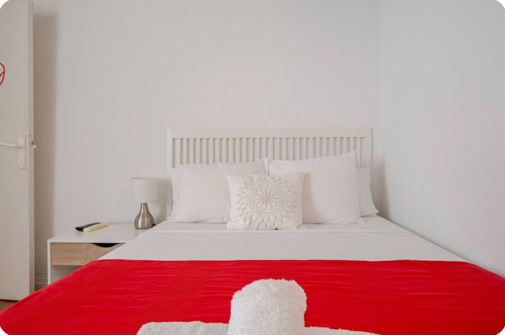 A neatly made bed with white bedding and a red blanket, a white bedside table with a lamp, and a white wall.