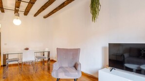 A modern living room with a grey armchair, a glass dining table with chairs, a flat-screen TV on a white stand, wooden beams on the ceiling, and a hanging plant. - The apartment in Opera Royal Palace! (Madrid)