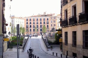 A street view with a building featuring multiple windows and balconies, a staircase leading to a higher level, and a sign indicating a location. - The apartment in Opera Royal Palace! (Madrid)