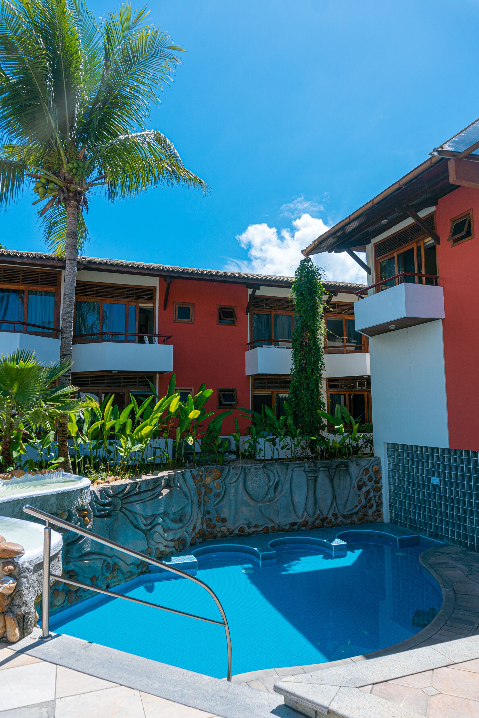 A pool area with a clear blue pool, surrounded by a stone wall and palm trees.