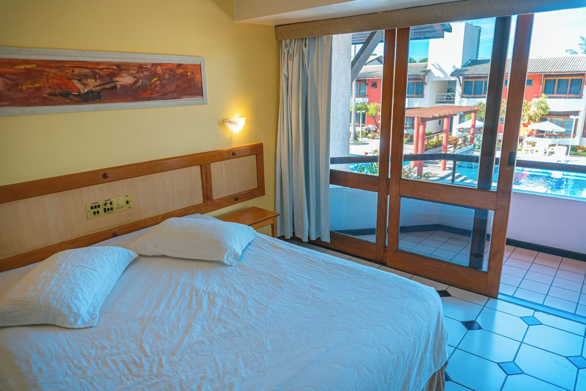 Habitación de hotel con cama, mesita de noche, lámpara de pared y un amplio ventanal con vistas a una piscina y edificios.