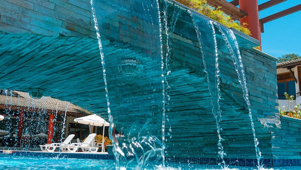 A pool with a waterfall feature, lounge chairs, and a covered area with a pergola. - Porto das Naus Praia Hotel (Porto Seguro)