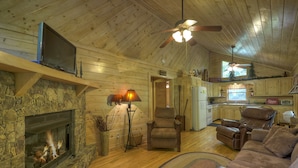 Een gezellig cabin interieur met een stenen open haard, houten wanden, een plafondventilator, een eethoek en een zithoek met een bank en fauteuils.