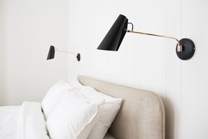 A bed with white linens and a beige headboard, two black wall-mounted lamps, and a white wall with vertical lines.